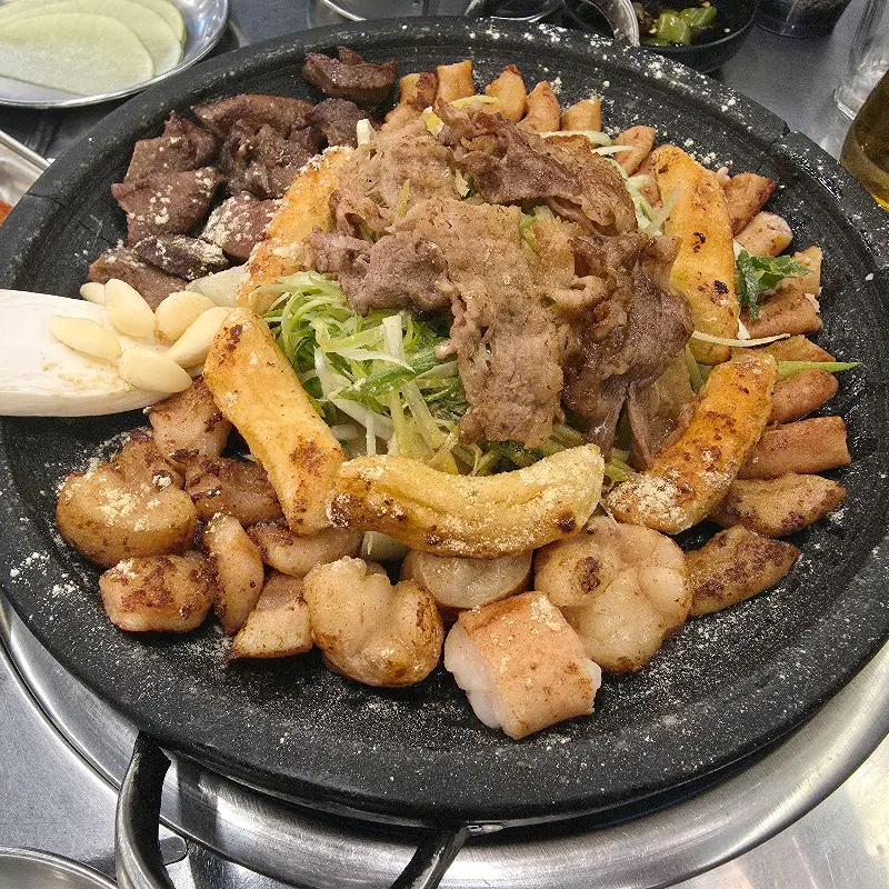 모듬 곱창이 돌판 위에 푸짐하게 담겨 있고, 곱창, 대창, 막창, 염통, 우삼겹, 떡, 양파, 마늘 등이 함께 구워져 있다.