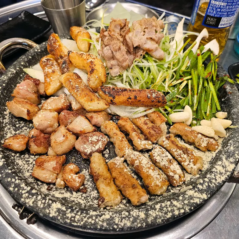 돌판 위에 곱창, 대창, 막창, 우삼겹, 떡, 양파, 마늘 등이 푸짐하게 구워져 있고, 깻잎 장아찌와 함께 먹는 모습이 보인다.