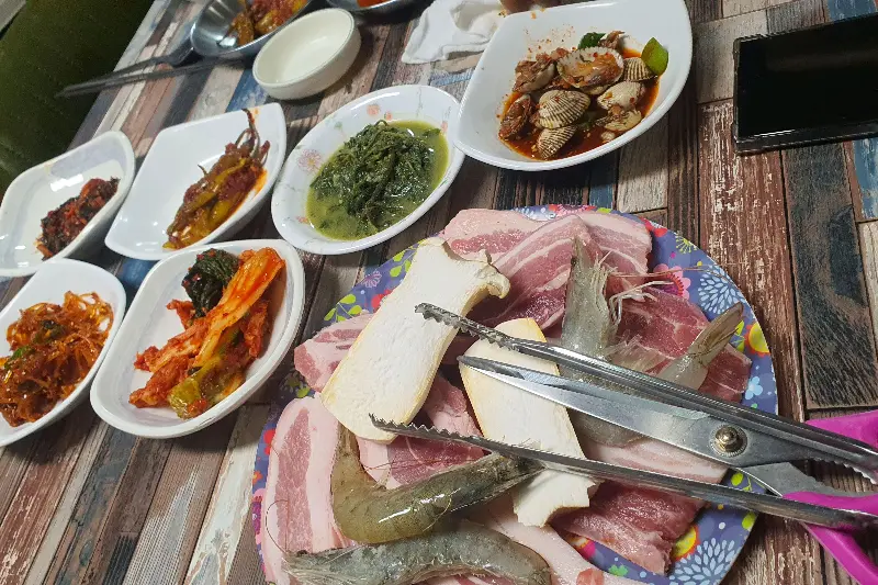 삼겹살, 버섯, 새우가 담긴 접시와 다양한 밑반찬이 차려진 테이블