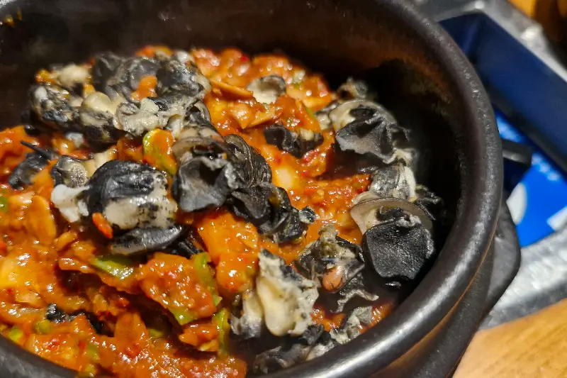 솥뚜껑 위에 삼겹살, 김치, 버섯, 양파, 마늘 등이 함께 구워지고 있는 모습