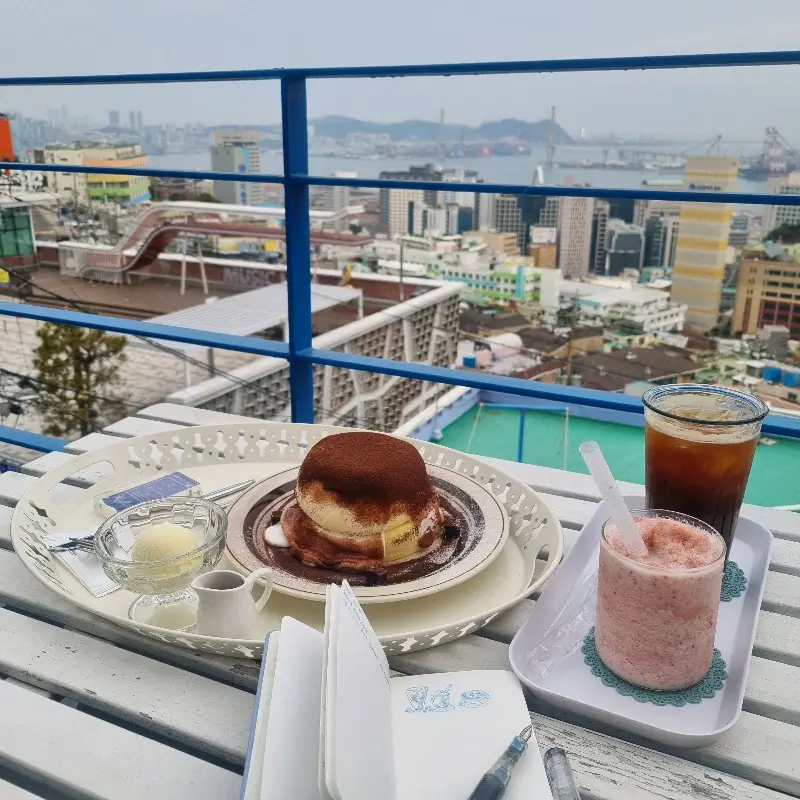 테이블에 놓인 티라미수 수플레, 딸기 스무디, 그리고 부산 시내 뷰를 담은 카페 파로스의 사진