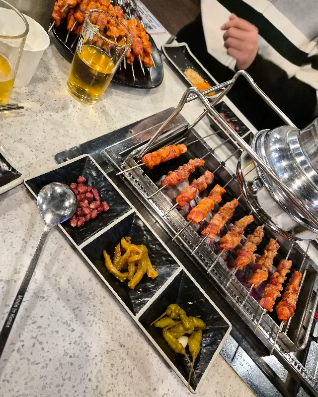 테이블 위에 놓인 양꼬치, 칭따오, 그리고 다양한 곁들임 찬들의 조화로운 모습