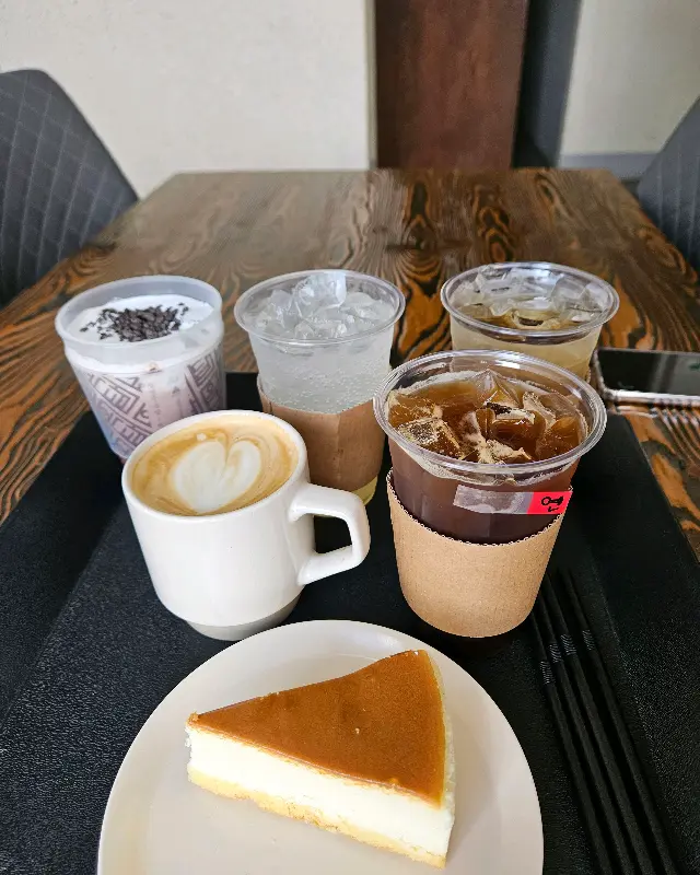 다양한 음료와 디저트가 테이블 위에 놓여져 있다. 딸기 우유, 라떼, 에이드, 그리고 조각 케이크가 보인다.