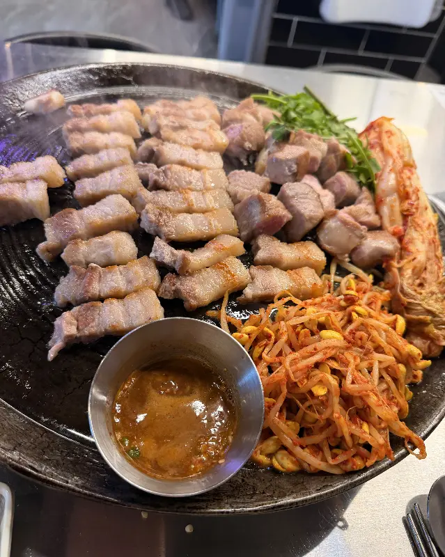 삼겹살, 김치, 콩나물, 미나리가 솥뚜껑에 구워진 모습