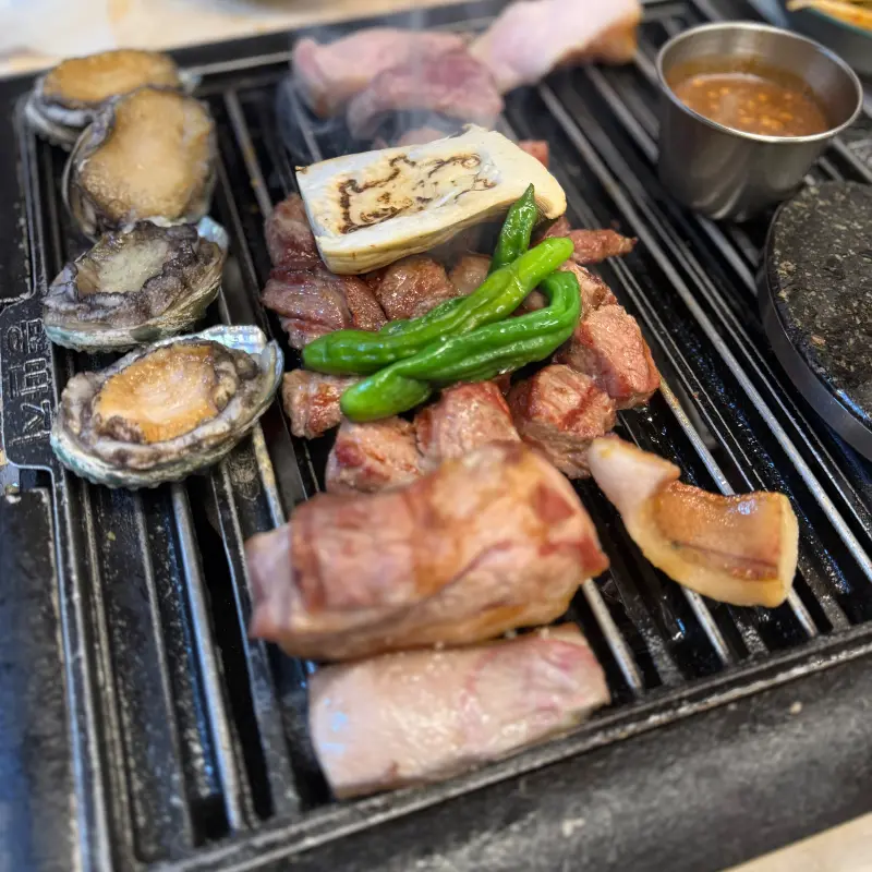 불판 위에서 맛있게 익어가는 흑돼지 근고기, 전복, 버섯, 꽈리꼬추의 클로즈업 모습