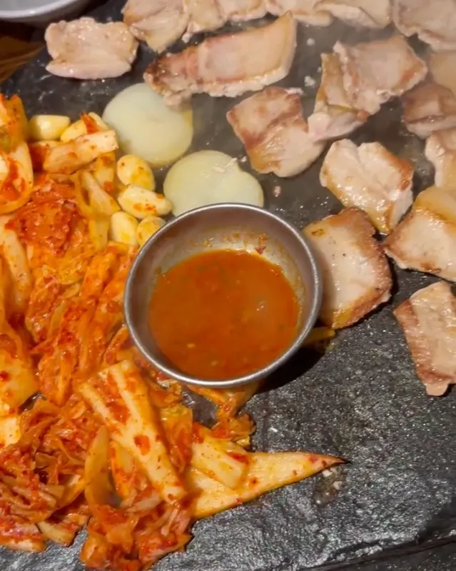 돌판 위에서 맛있게 익어가는 삼겹살과 김치, 그리고 가운데 놓인 소스