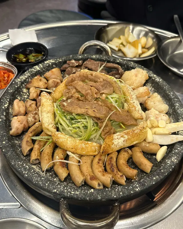 모듬 곱창이 돌판 위에 가지런히 놓여 있다. 곱창, 대창, 막창, 염통, 우삼겹, 떡, 양파, 마늘 등이 함께 구워져 있고, 곱창 위에는 하얀 치즈 가루가 뿌려져 있다.