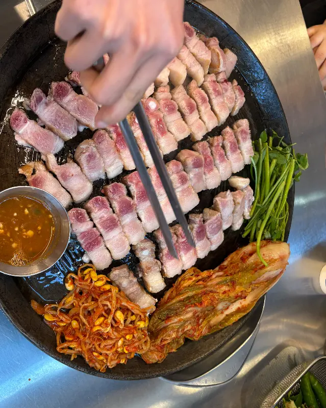 솥뚜껑에 구워진 삼겹살, 김치, 콩나물, 미나리의 모습