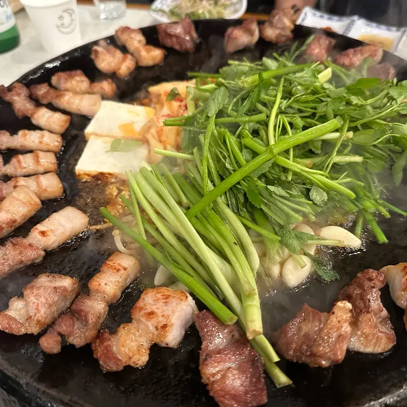 무쇠판에 구워진 삼겹살, 김치, 미나리의 모습