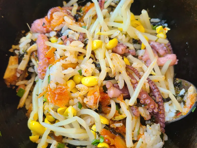 바지락돌솥비빔밥을 숟가락으로 크게 떠올린 모습. 밥알과 재료들이 조화롭게 어우러져 먹음직스럽다.