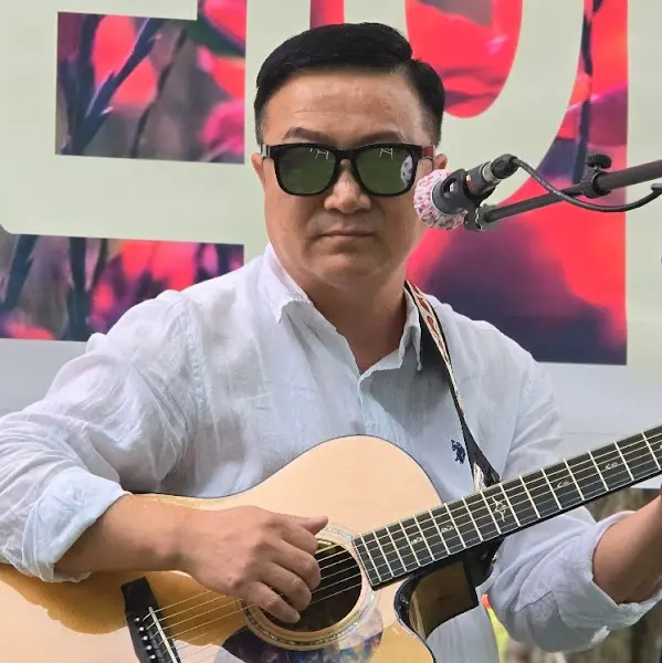 기타를 연주하는 남성의 모습. 그는 선글라스를 끼고 있으며, 흰색 셔츠를 입고 있다.