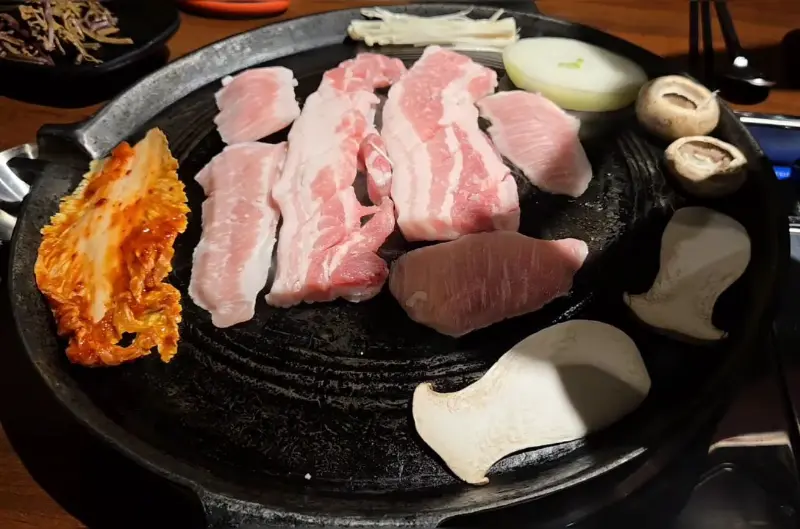 솥뚜껑 한가득 구워진 삼겹살, 김치, 버섯, 콩나물