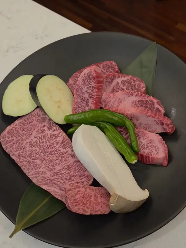 검은 접시에 담긴 신선한 한우는 붉은색 마블링이 선명하게 드러나고, 꽈리고추, 새송이버섯, 가지가 함께 놓여 있어 먹음직스러운 플레이팅을 완성합니다.