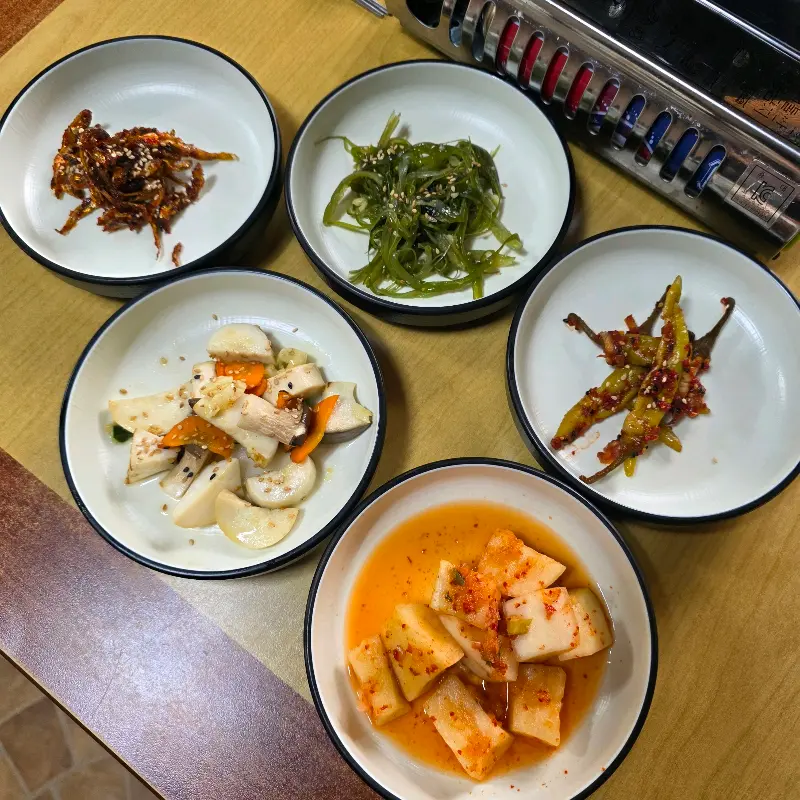 다섯 종류의 반찬이 각각 작은 접시에 담겨 있다. 볶음김치, 해초무침, 무생채 등이 보인다.