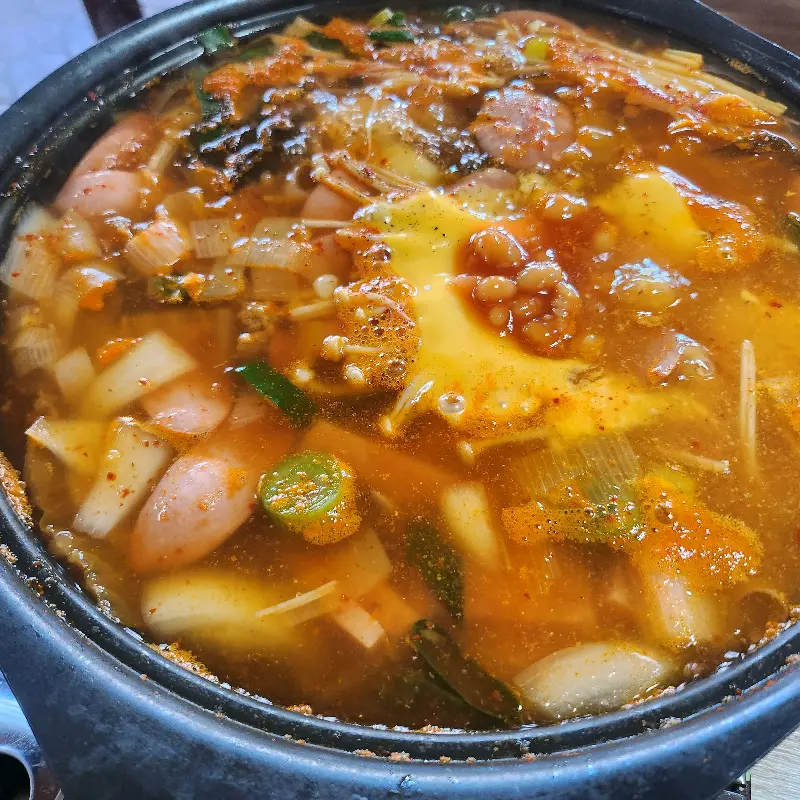 보글보글 끓고 있는 부대찌개의 모습이 담겨있다. 햄, 소시지, 야채, 김치 등이 푸짐하게 들어가 있고, 체다치즈 한 장이 녹아내리고 있다.