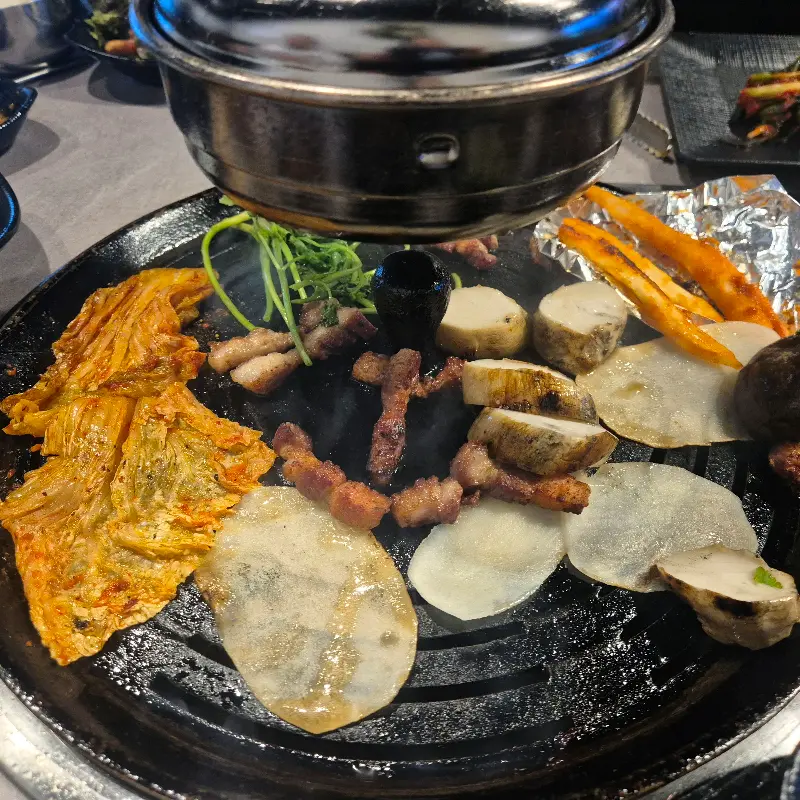 불판 위에서 익어가는 삼겹살, 김치, 버섯, 떡