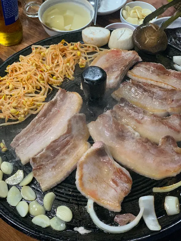 솥뚜껑 위에 삼겹살, 김치, 콩나물, 버섯, 양파가 함께 구워지는 모습