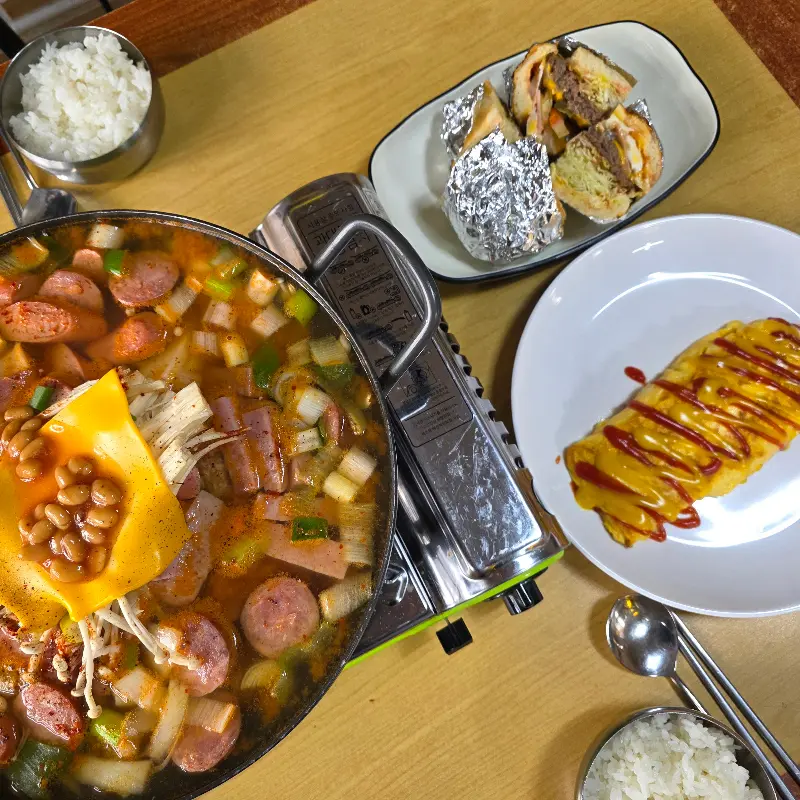 테이블 위에 부대찌개, 계란말이, 햄버거, 밥 등이 놓여 있는 전체적인 모습이 담겨 있다.