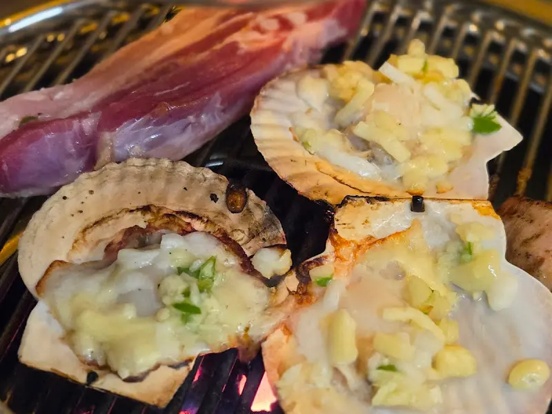 연탄불 위에서 구워지는 삼겹살과 가리비. 고기의 표면은 갈색으로 맛있게 익어가고, 가리비는 치즈와 함께 녹아내리고 있다.