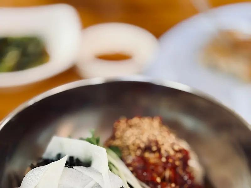 스테인리스 그릇에 담긴 막국수 위로 김가루, 무순, 채 썬 백김치가 올려져 있고, 붉은 양념장이 한가운데 놓여 있다.