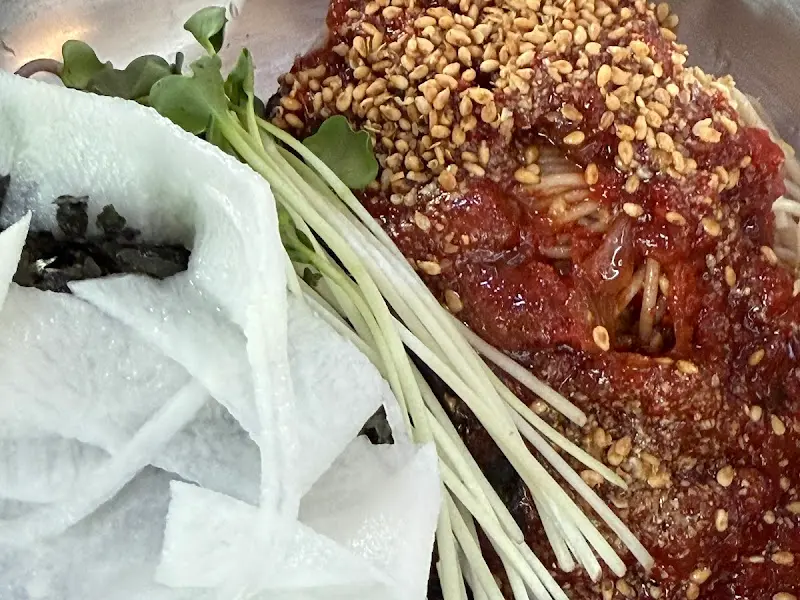 막국수 위에 김가루, 무순, 채 썬 백김치가 넉넉하게 올려져 있고, 붉은 양념장이 면을 덮고 있다. 참깨가 듬뿍 뿌려져 있어 고소한 풍미가 느껴진다.