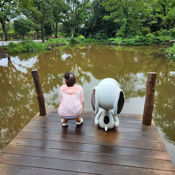 나무 데크 위에 여자아이와 스누피 캐릭터 인형이 연못을 바라보고 있는 모습