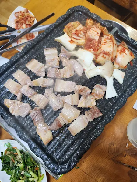 불판 위에 삼겹살과 김치가 구워지고 있고, 주변에 여러 반찬 그릇들이 놓여 있는 모습