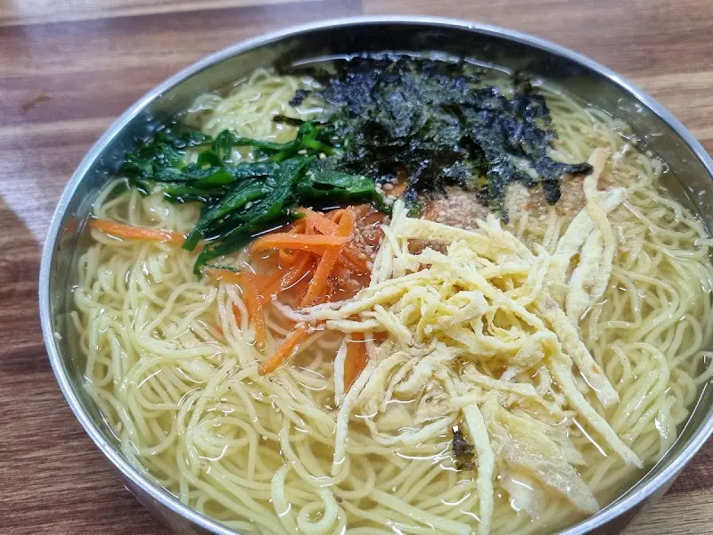 스테인리스 그릇에 담긴 잔치국수의 클로즈업 사진. 면 위에는 김 가루, 채 썬 당근, 계란 지단, 애호박이 고명으로 올려져 있다.