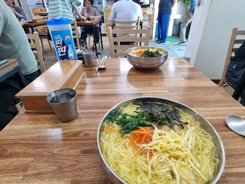 테이블에 놓인 스테인리스 그릇에 담긴 잔치국수 사진. 테이블은 나무 재질로 되어 있으며, 냅킨 케이스와 물통이 함께 놓여 있다.