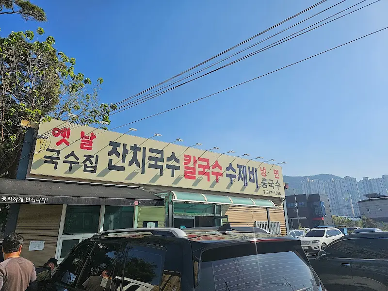 식당 건물 외부 사진. '옛날 잔치국수 칼국수 수제비'라고 적힌 간판이 달려 있으며, 건물 앞에는 차들이 주차되어 있다.