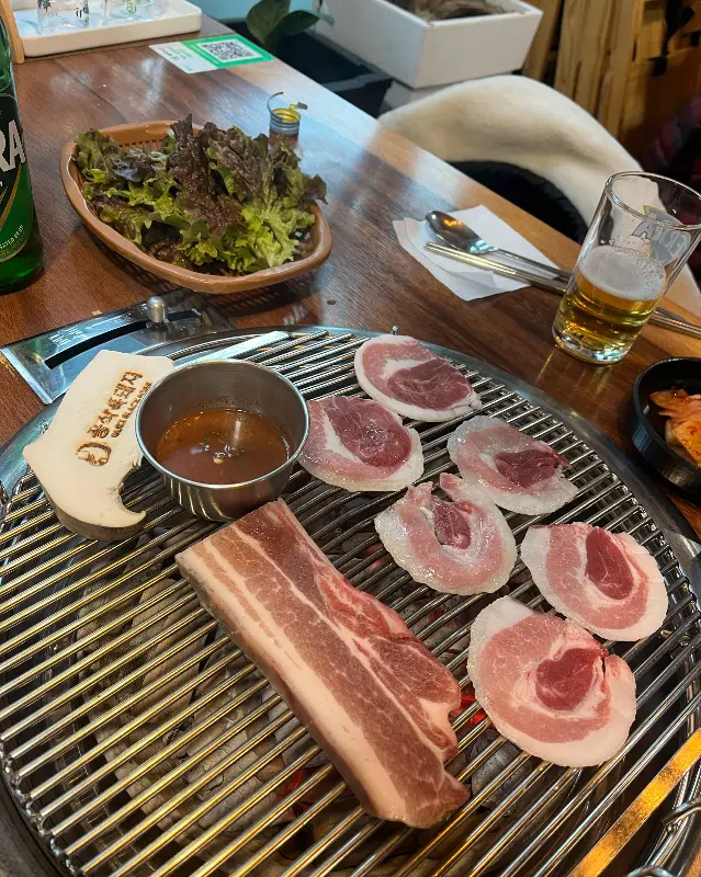 불판 위에 흑돼지 오겹살이 구워지고 있다. 멜젓과 쌈 채소, 맥주가 함께 놓여 있다.