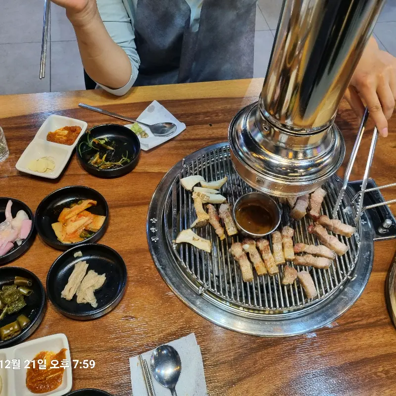 테이블 위에서 직원분이 흑돼지 오겹살을 굽고 있다. 다양한 밑반찬과 소스들이 함께 놓여 있다.