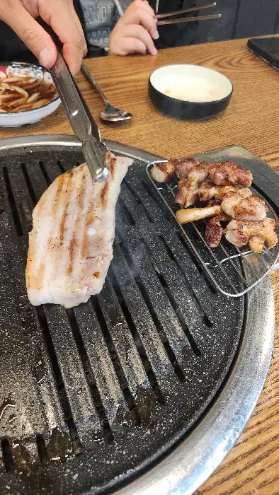 집게로 삼겹살 한 점을 불판에 올리며 지글거리는 소리가 나는 모습. 옆에는 잘 구워진 고기들이 빼곡히 놓여 있다.
