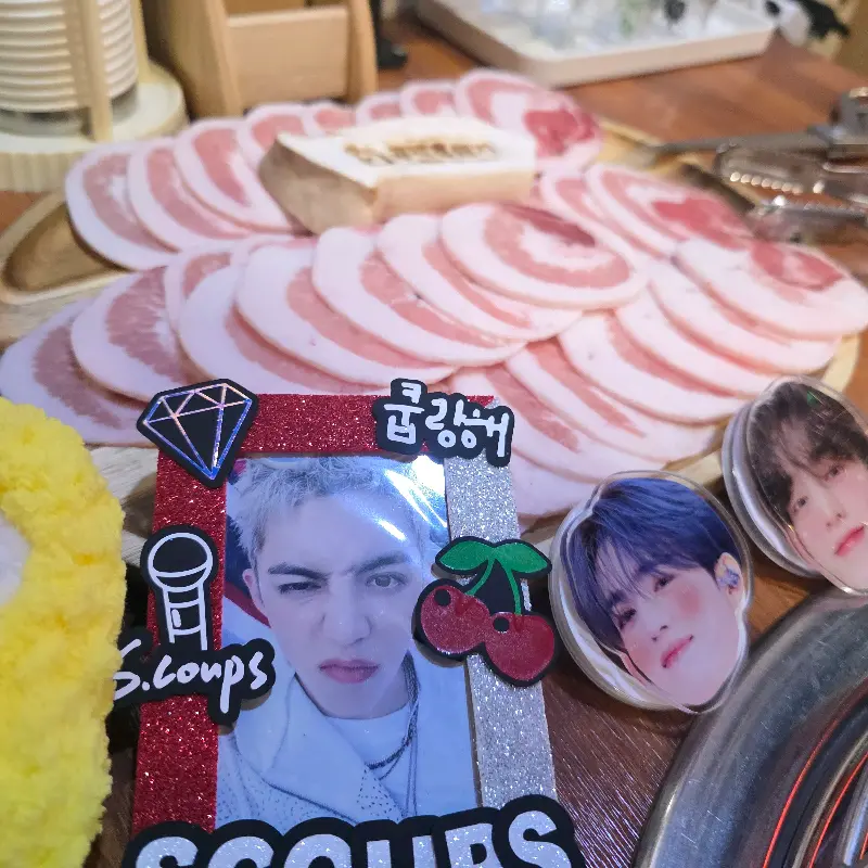 테이블 위에 흑돼지 오겹살이 가득 담긴 접시와 함께 귀여운 캐릭터 인형과 사진 프레임이 놓여 있다.