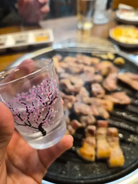 작은 잔에 담긴 술과 함께 불판 위에서 구워지고 있는 고기들의 모습. 따뜻한 조명 아래 낭만적인 분위기를 연출한다.