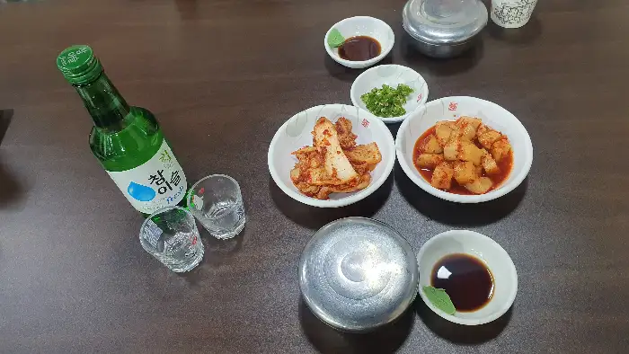 테이블 위에 놓인 소주병, 잔, 김치, 깍두기, 밥그릇, 소스 그릇