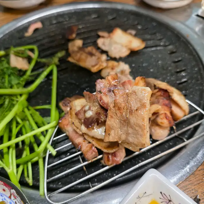 잘 구워진 삼겹살과 신선한 미나리가 함께 불판 위에 놓여 있다. 미나리의 푸른색과 삼겹살의 갈색 빛깔이 대조를 이룬다.