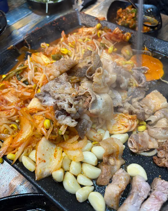 불판 위에서 지글지글 익어가는 대패삼겹살과 김치, 콩나물, 마늘