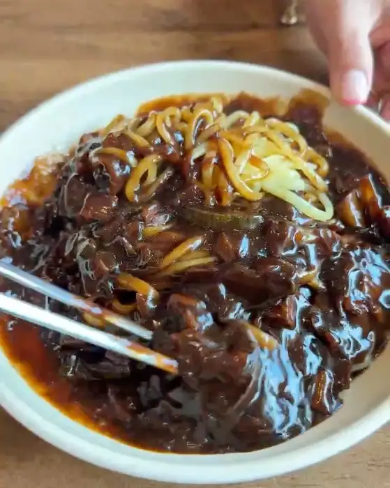 검은색 짜장 소스가 듬뿍 부어진 면 위에 채 썬 노란색 단무지와 함께 젓가락으로 면을 들어 올리는 모습.