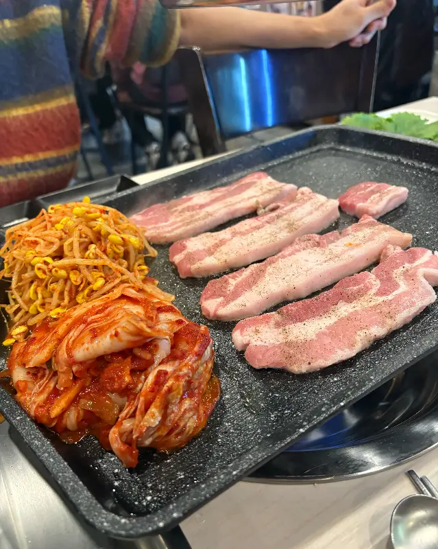 불판 위에서 노릇하게 익어가는 삼겹살과 김치, 콩나물