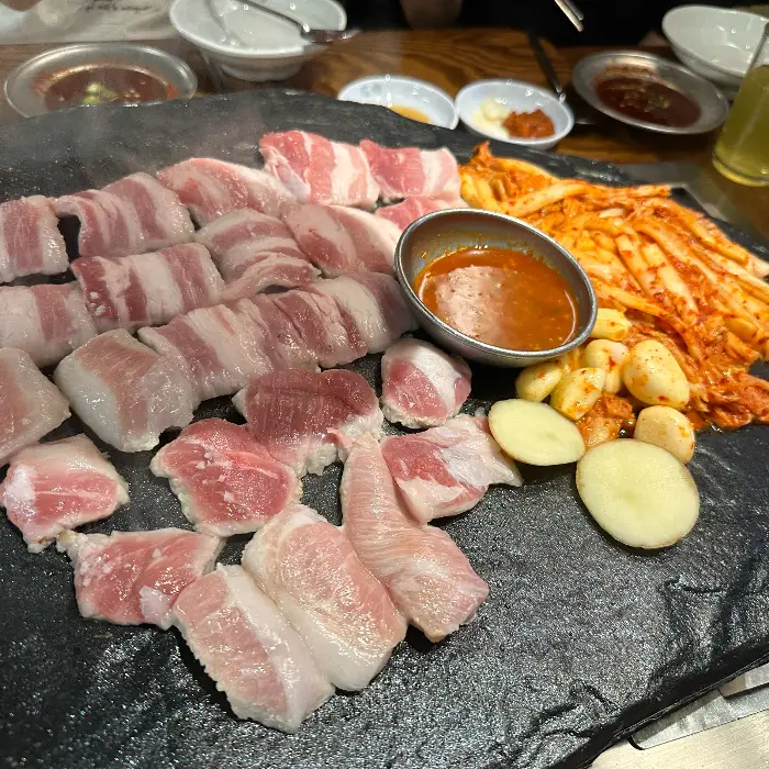 돌판 위에 먹음직스럽게 구워진 삼겹살과 곁들임 찬이 놓여 있는 모습