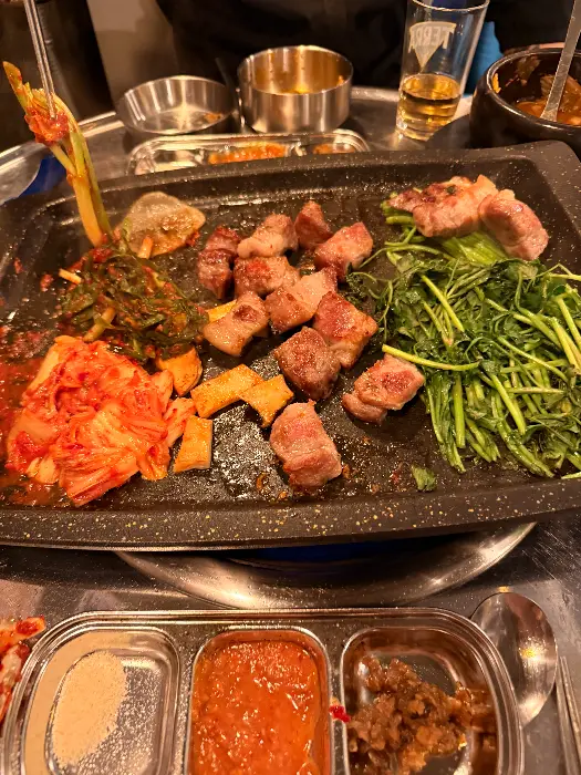 고기, 김치, 미나리, 깻잎 등 다양한 반찬과 함께 불판 위에서 익고 있는 고기