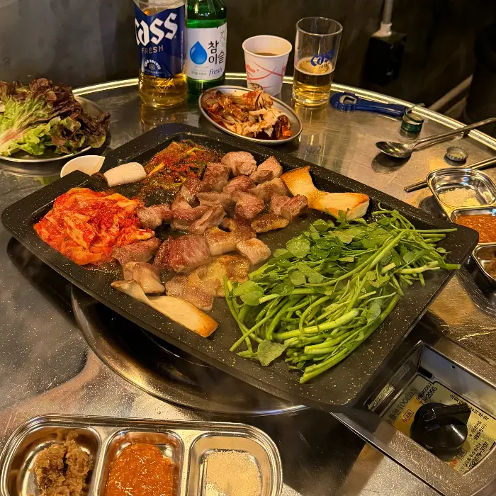 불판 위에 삼겹살, 김치, 미나리, 깻잎 등이 함께 구워지고 있고, 테이블 위에는 맥주와 소주가 놓여 있습니다