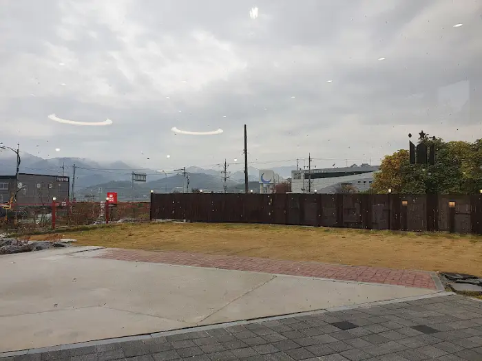 View of a paved area with some grass and a wooden fence, with distant hills under a cloudy sky