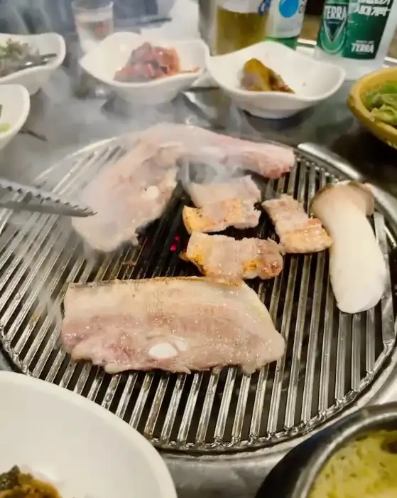 지글지글 숯불 위에서 익어가고 있는 흑돼지 삼겹살과 버섯, 마늘. 김이 모락모락 피어오르고 있다.