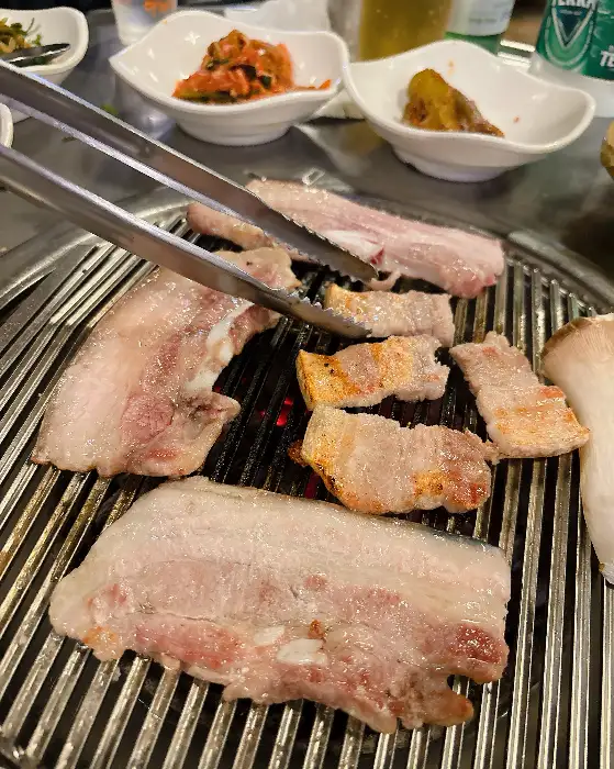 집게로 흑돼지 삼겹살을 집어 숯불 위로 옮기는 모습. 다른 조각들도 불판 위에서 익어가고 있다.