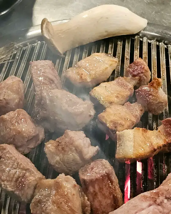 숯불 위에서 익어가는 흑돼지 고기와 버섯. 흰색 버섯의 갓 부분이 먹음직스럽게 구워지고 있다.