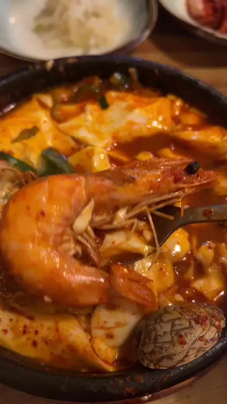 해물 순두부 찌개가 담긴 검은 뚝배기 클로즈업 사진. 새우와 조개, 순두부가 보인다.