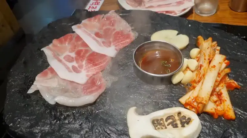 돌판 위에 김치, 마늘, 버섯과 함께 삼겹살이 구워지는 모습