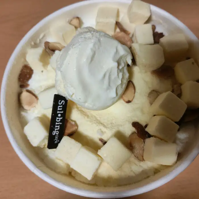 치즈 빙수. 뽀얀 얼음 위에 큐브 모양의 치즈와 아몬드 슬라이스가 올라가 있고, 'Sul-bing' 로고 플레이트가 꽂혀 있습니다.
